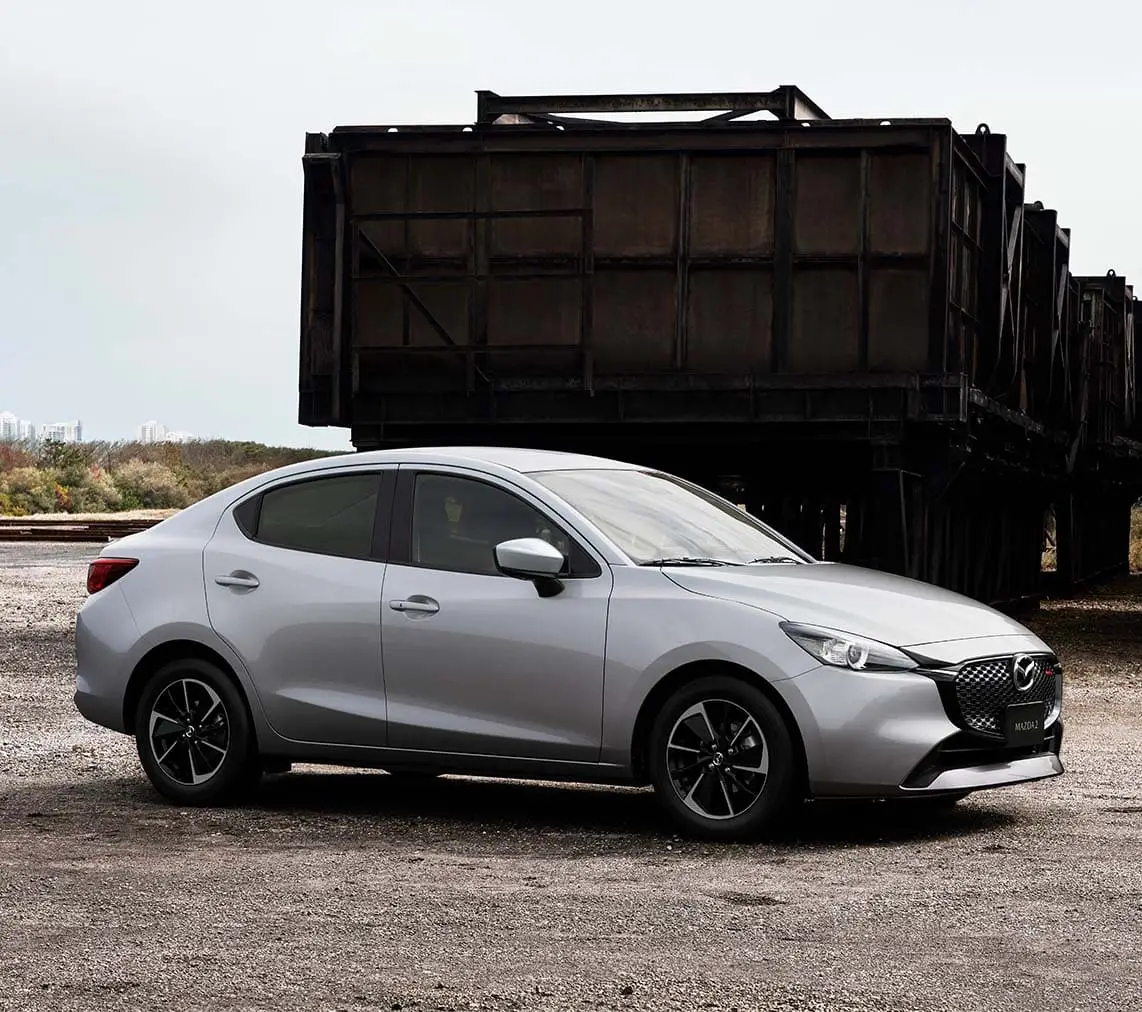 Mazda 2 Sedán color Gris Aéreo, vista lateral en entorno industrial